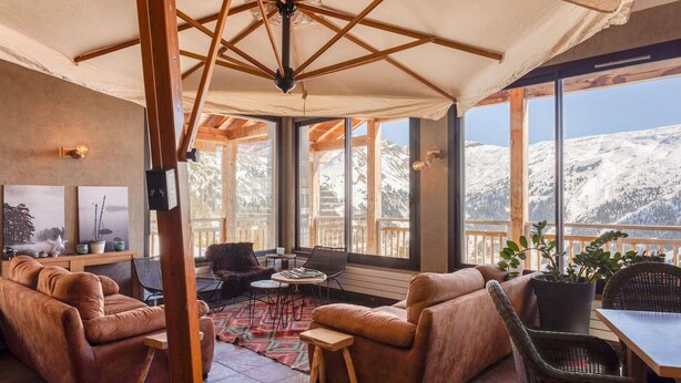 A cozy lounge area with leather sofas, a wooden table, and a view of snow-covered mountains through large windows. - Cozy Mountain Stay w/ Balcony (Arâches-La-Frasse)