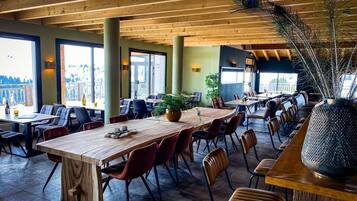 A spacious indoor dining area with wooden tables and chairs, large windows, and a decorative plant centerpiece.