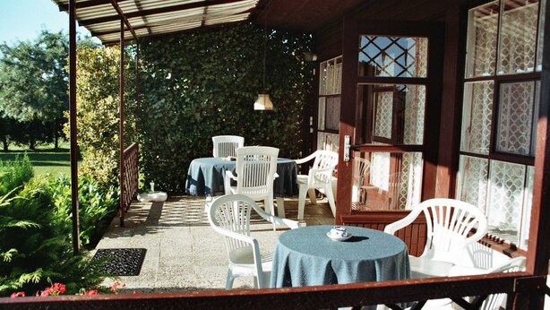 A covered patio area with white chairs and tables, surrounded by a garden and a wooden building with patterned windows. - Ferienhaus Schmidt, Erika - Ferienhaus 7 (Mönkebude)