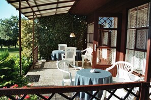 A covered patio area with white chairs and tables, surrounded by a garden and a wooden building with patterned windows. - Ferienhaus Schmidt, Erika - Ferienhaus 7 (Mönkebude)
