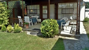 A covered outdoor seating area with tables and chairs, surrounded by greenery and a small garden. - Ferienhaus Schmidt, Erika - Ferienhaus 7 (Mönkebude)