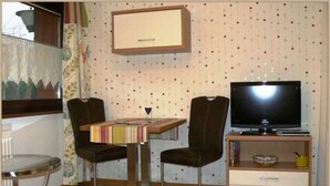 Une petite pièce avec un téléviseur, une table avec des chaises, un petit meuble et une fenêtre avec des rideaux.