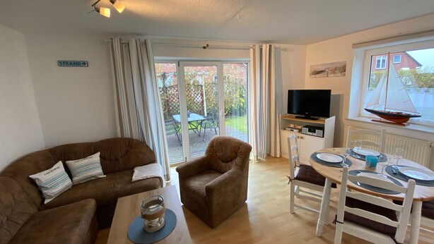 A cozy living room with a sofa, armchair, and a dining table set for two. A television is mounted on the wall, and there's a window with a sailboat model on the sill.