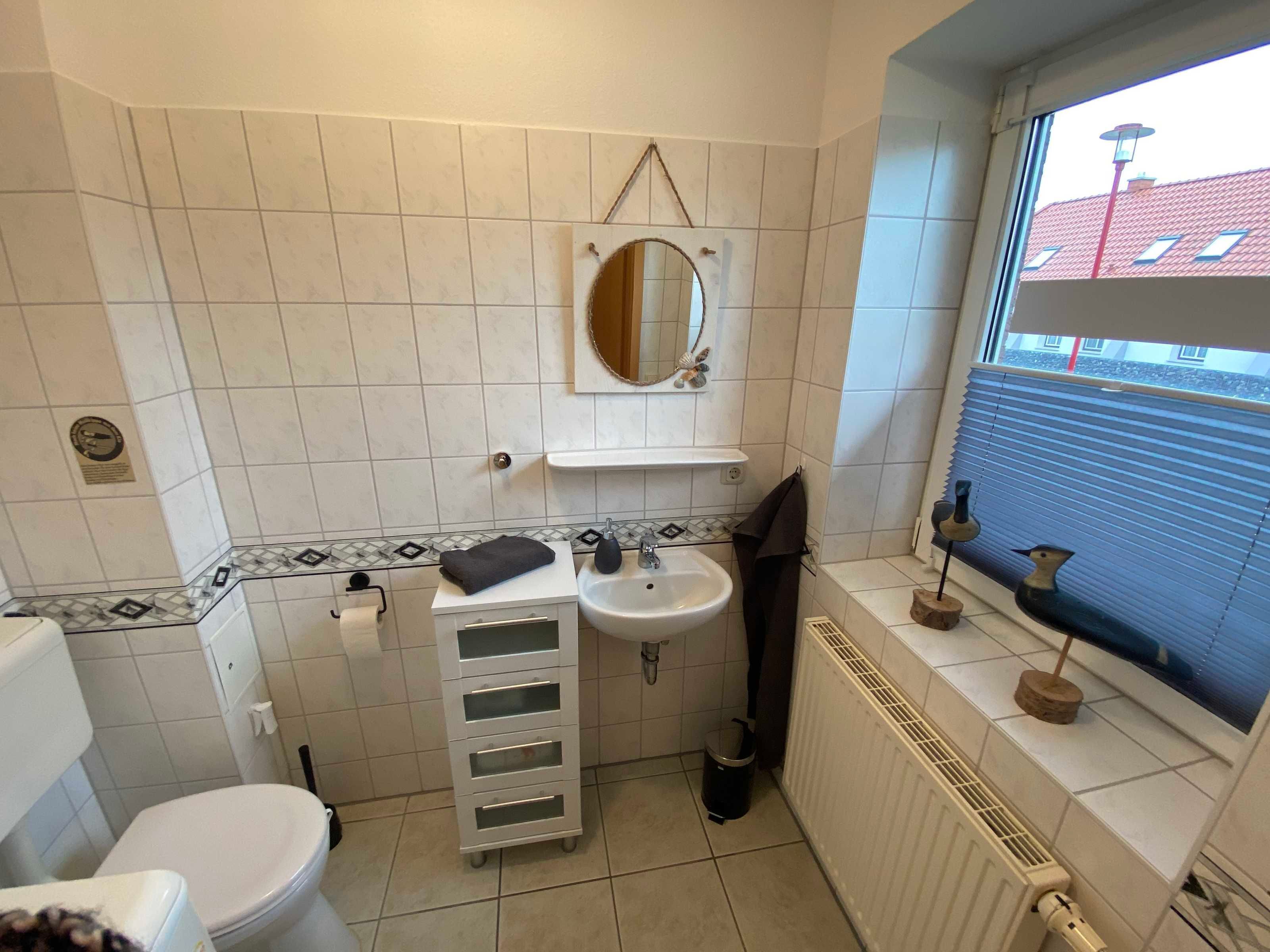 A small bathroom with a toilet, sink, and mirror. There is a towel rack, a small shelf, and a window with blinds.