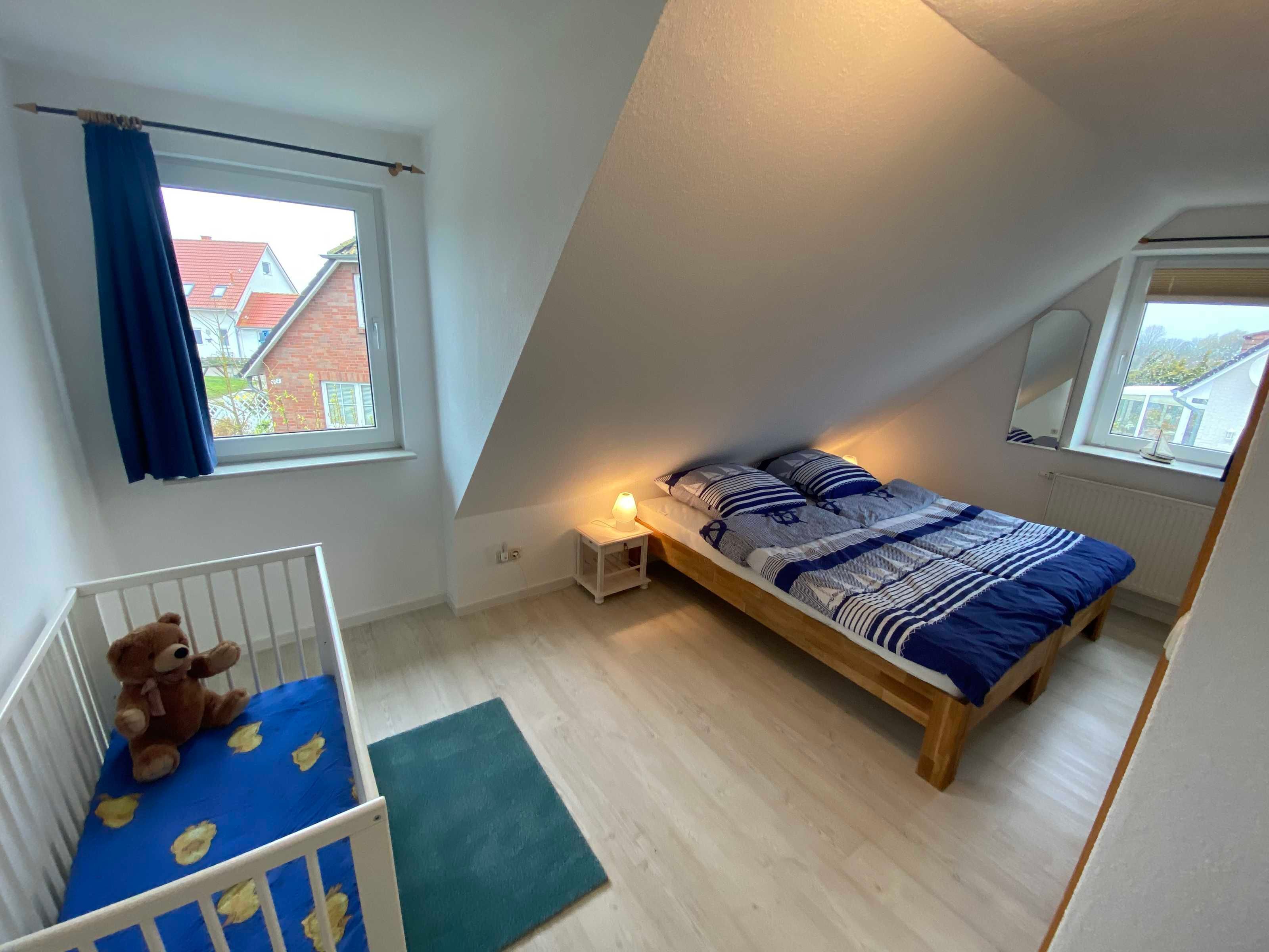 A bedroom with a sloped ceiling, a bed with a blue and white striped bedspread, a crib with a teddy bear, and a window with a view of houses.
