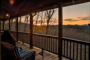 A covered outdoor area with a wooden deck, a bench, and a view of a sunset over a landscape with trees and hills.