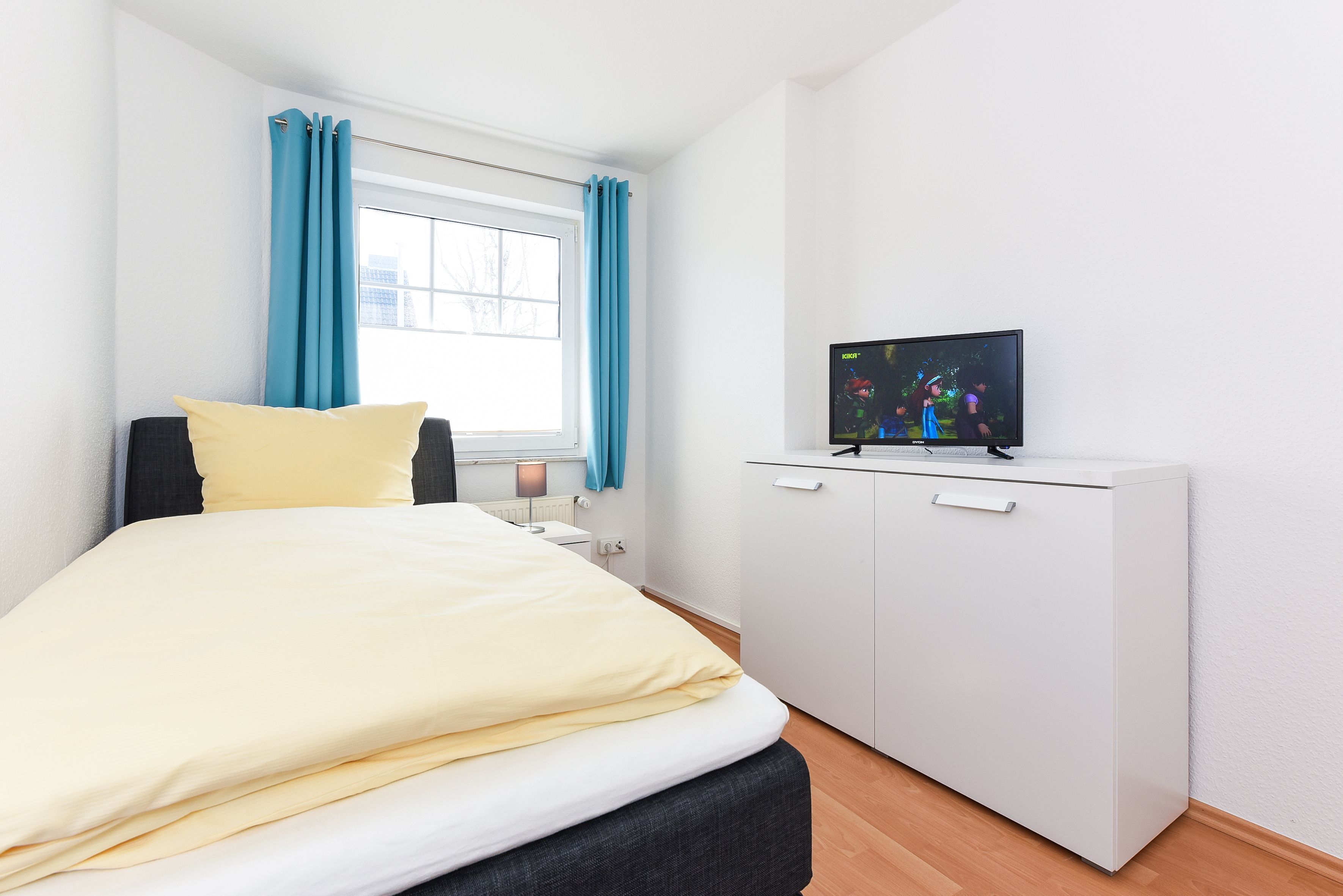 A bedroom with a bed, a TV on a white cabinet, a window with curtains, and a nightstand with a lamp.