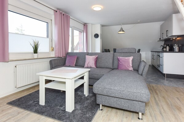 A modern living room with a grey sectional sofa, a white coffee table, and a kitchen area with white cabinets and appliances.