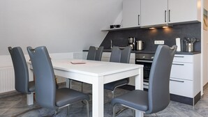A modern kitchen with white cabinets, a white countertop, and a dining area with a white table and grey chairs. - Ferienwohnung Brise - Mein Urlaubshaus (Esens)