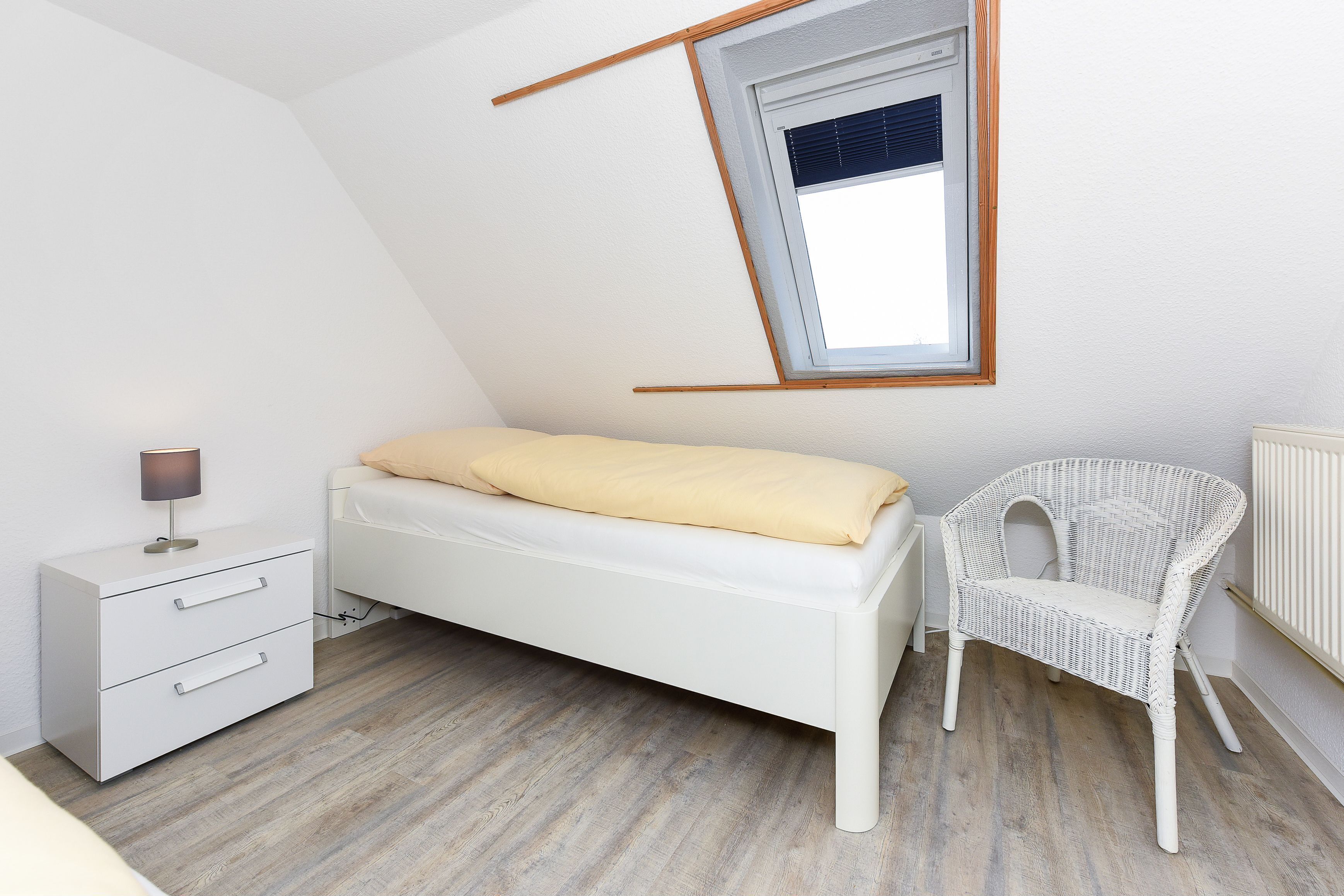 A bedroom with a white bed, a white nightstand, a wicker chair, and a window with blinds.
