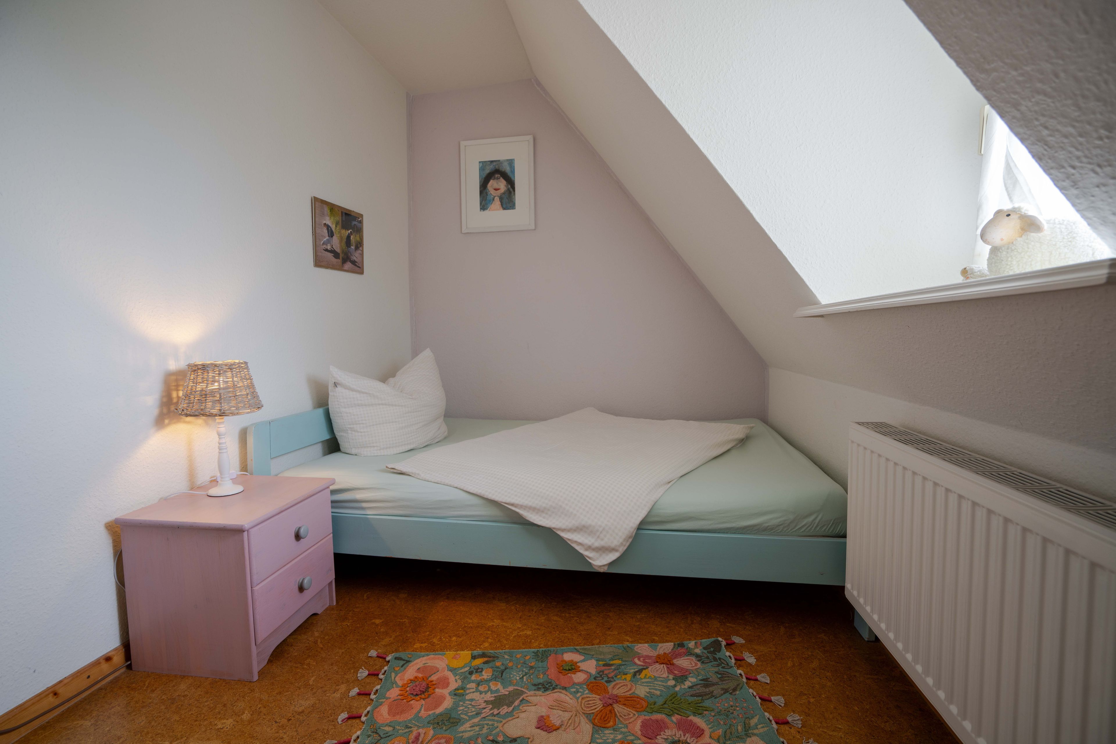 Een slaapkamer met een schuine kap, een bed met een witte deken, een nachtkastje met een lamp en een radiator.