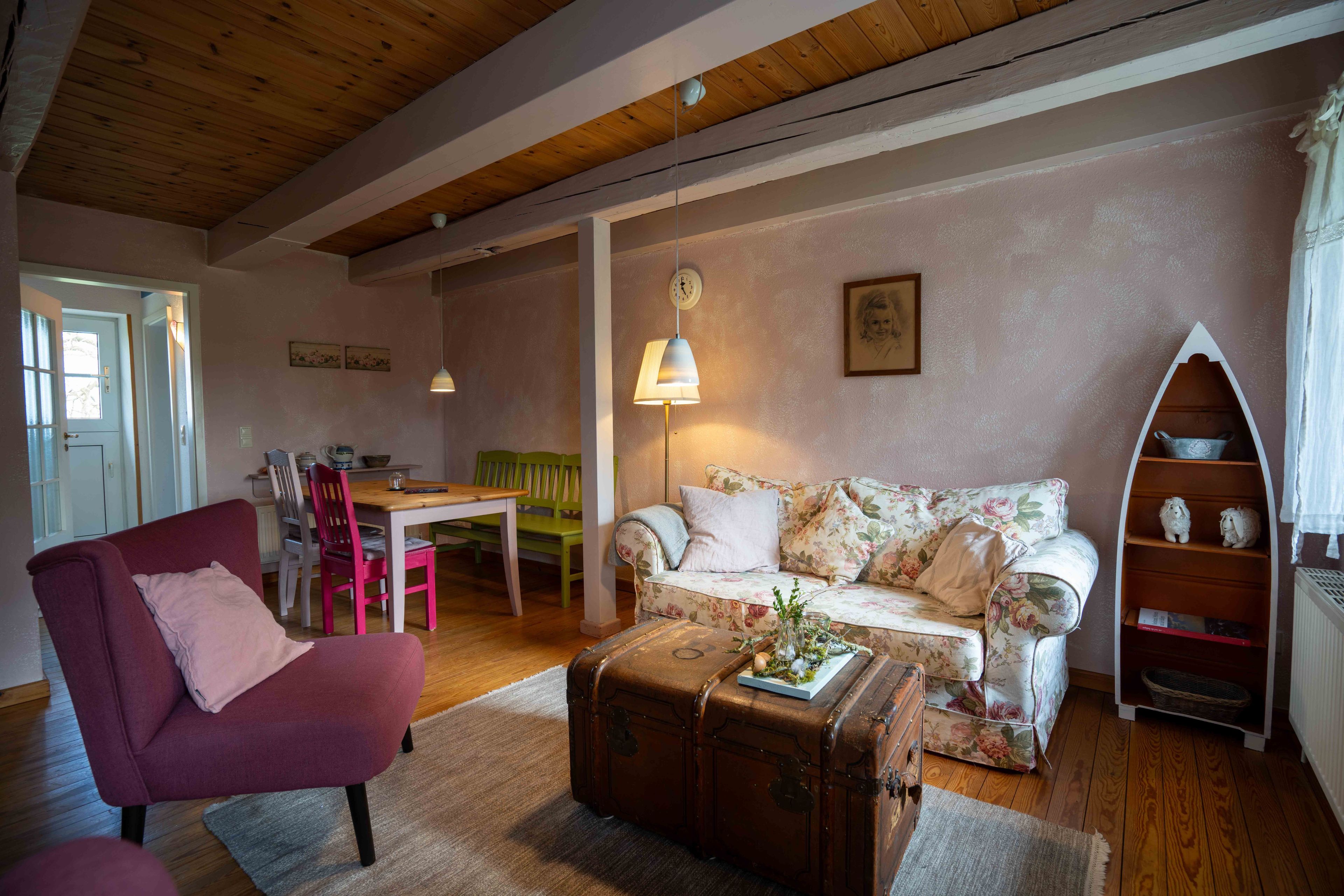 Een gezellige woonkamer met een bloemenbank, een rode fauteuil, een houten eettafel en een vintage koffer als salontafel.