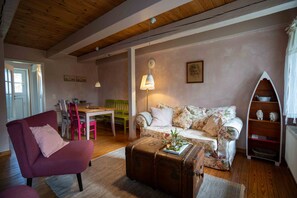 Un salon chaleureux avec un canapé à motifs floraux, un fauteuil rouge, une table à manger en bois et une valise ancienne servant de table basse.