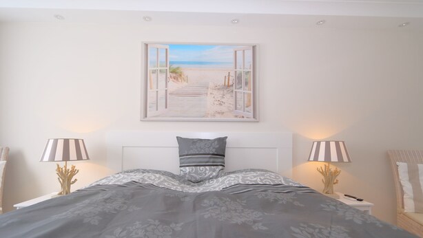 Een slaapkamer met een bed, twee bedlampen, een ingelijst strandfoto en een wit hoofdeinde.