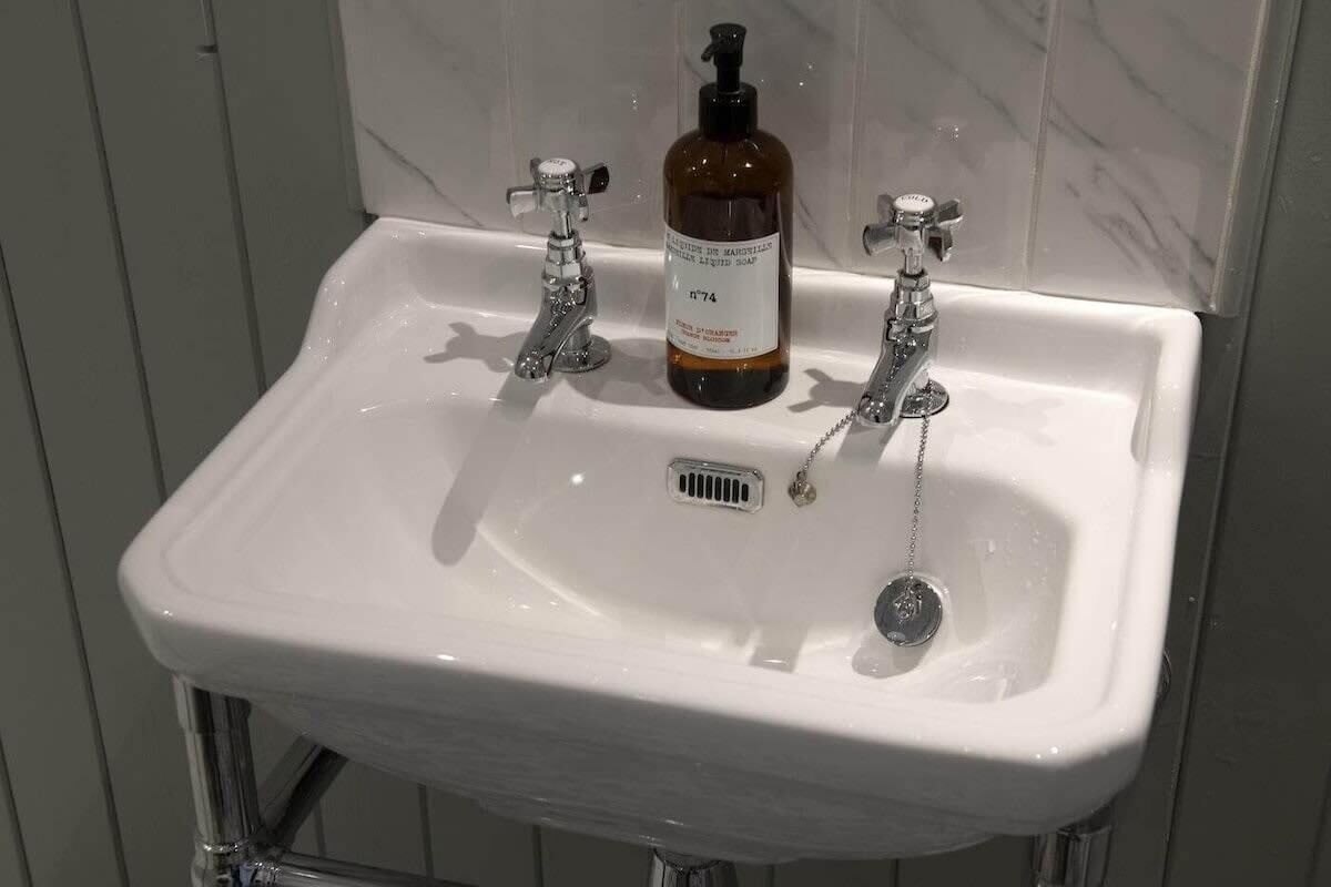 A white bathroom sink with two chrome faucets and a bottle of liquid soap.