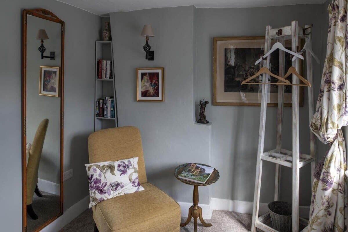 A room with a beige armchair, a small side table, a mirror, a bookshelf, a coat rack, and a framed picture on the wall.