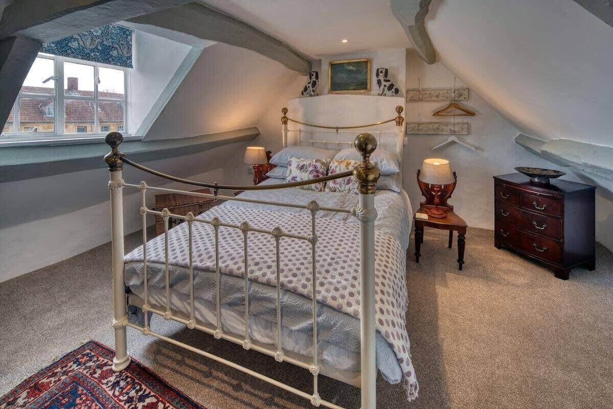 Een slaapkamer met een bed met een wit metalen frame, een groot raam, een houten dressoir en een kamerkleed met een patroon.
