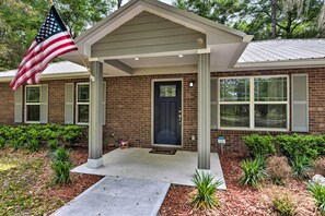 A brick building with a dark blue door, a covered porch, and an American flag. - Serene Live Oak Home w/ Fire Pit by River + SOSMP! (Live Oak)