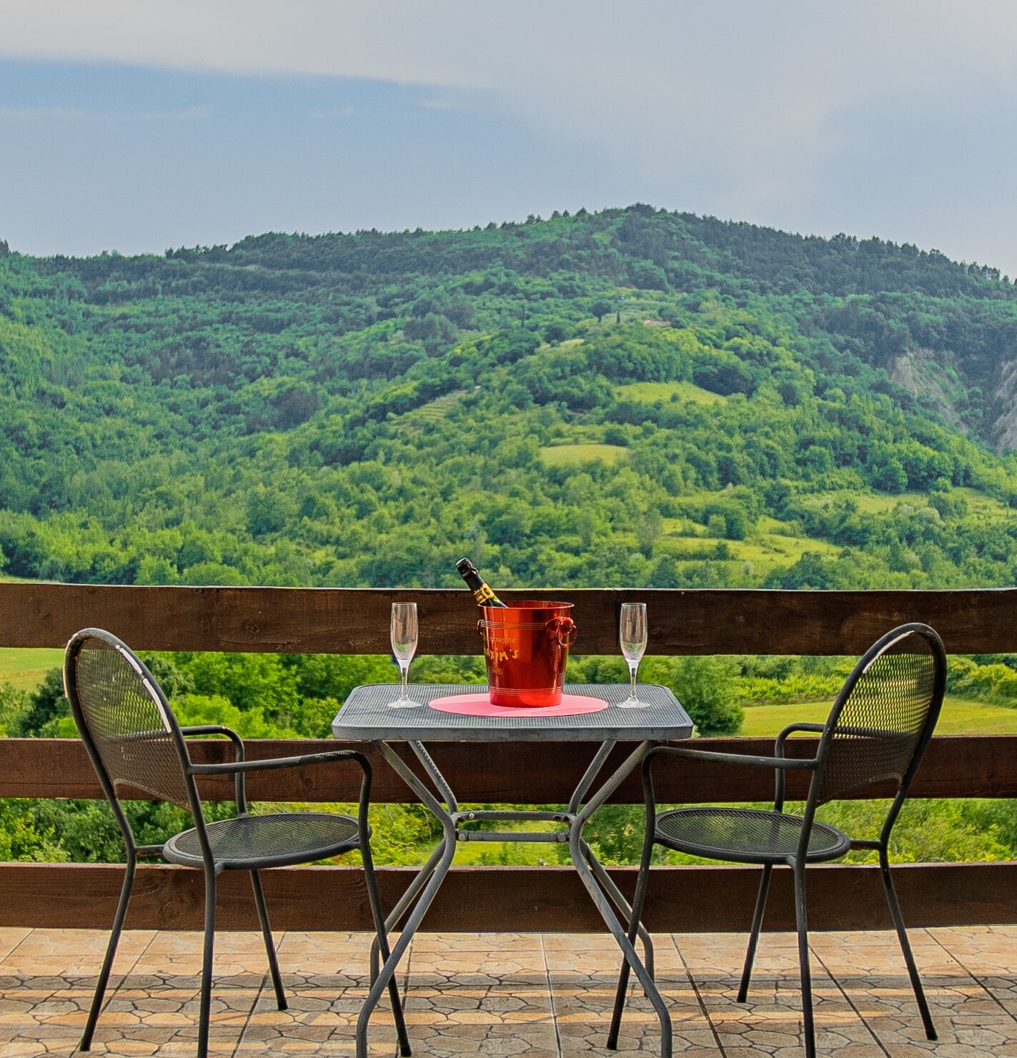 Una mesa con dos sillas y un balde rojo en un balcón con vistas a un valle verde exuberante.