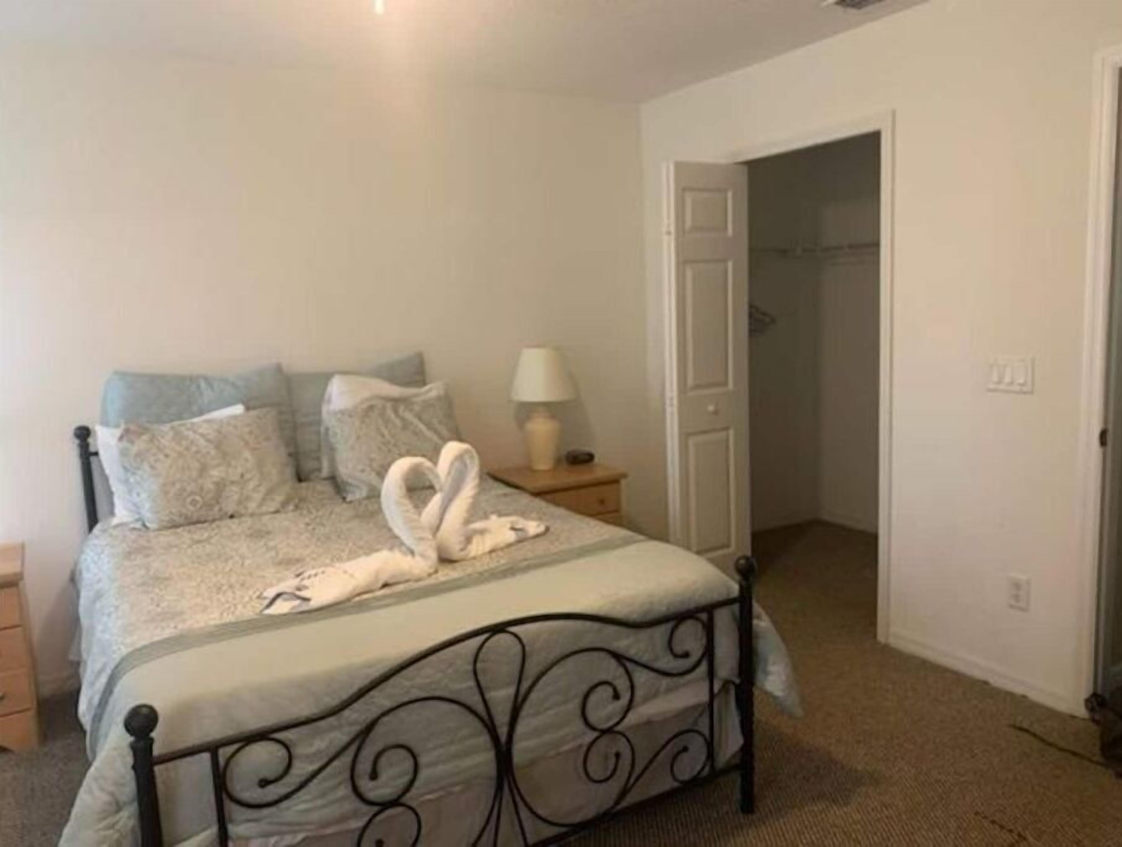 Quarto com cama, almofadas, mesa de cabeceira com abajur e porta para outro cômodo.