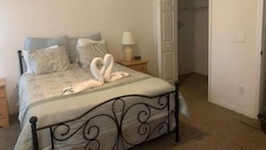 Quarto com cama, almofadas, mesa de cabeceira com abajur e porta para outro cômodo.