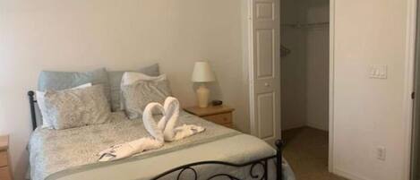 Quarto com cama, almofadas, mesa de cabeceira com abajur e porta para outro cômodo.