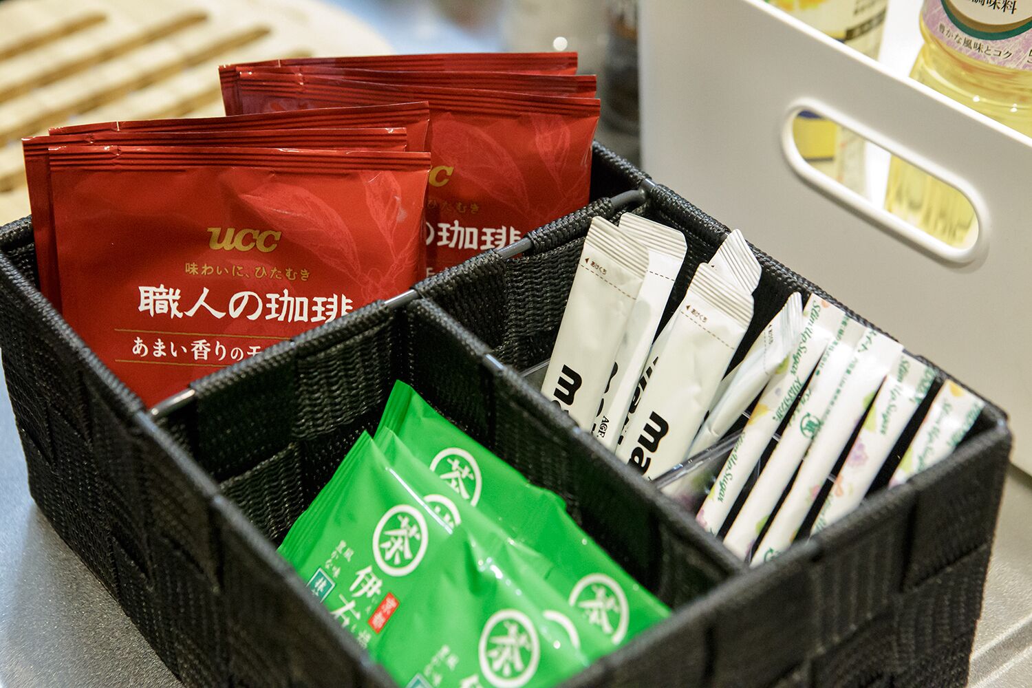 A black basket containing red and green packets, some white packets, and a white container with a yellow liquid.