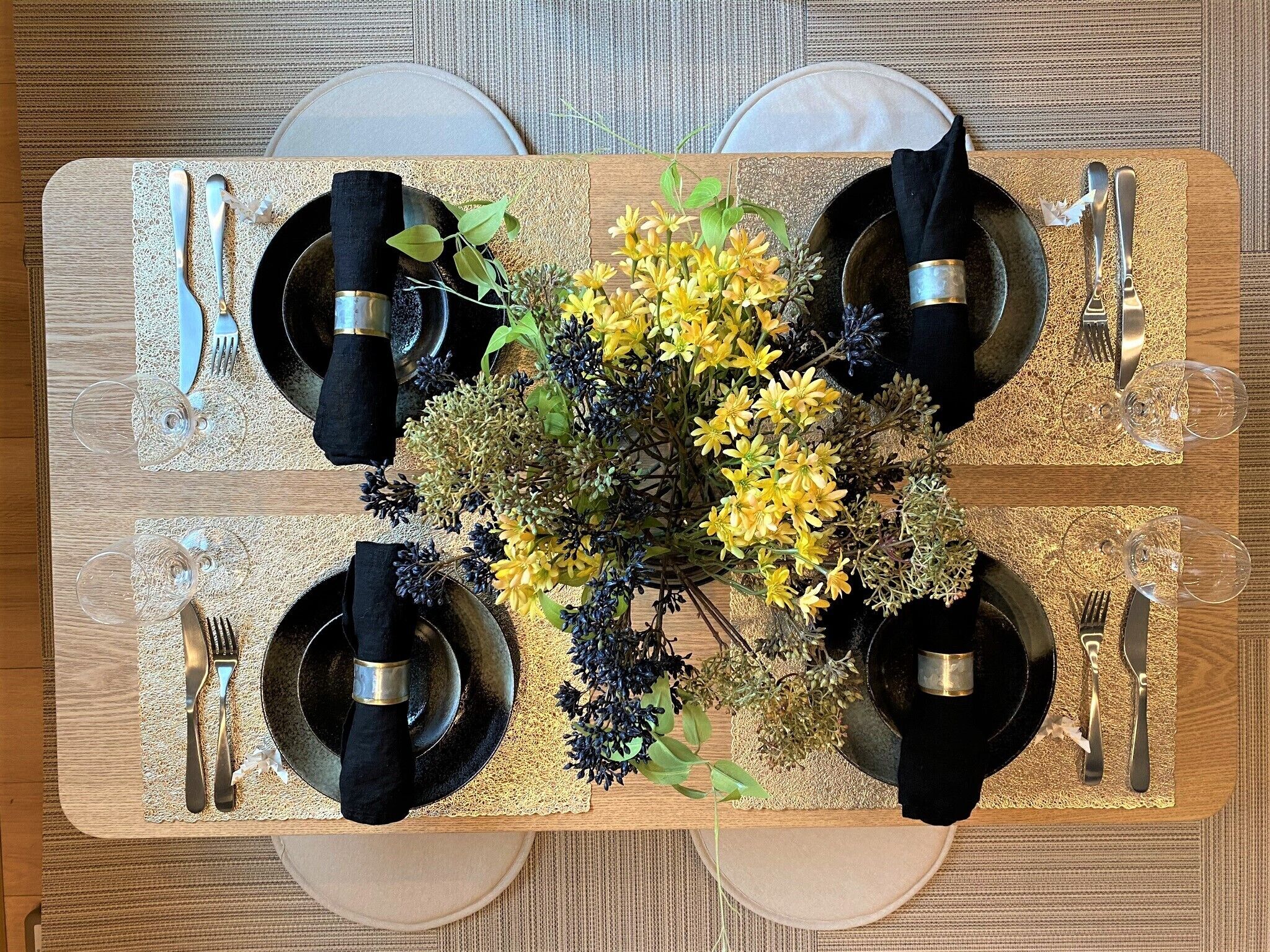 A rectangular dining table with four black plates, silverware, and a centerpiece of yellow flowers.