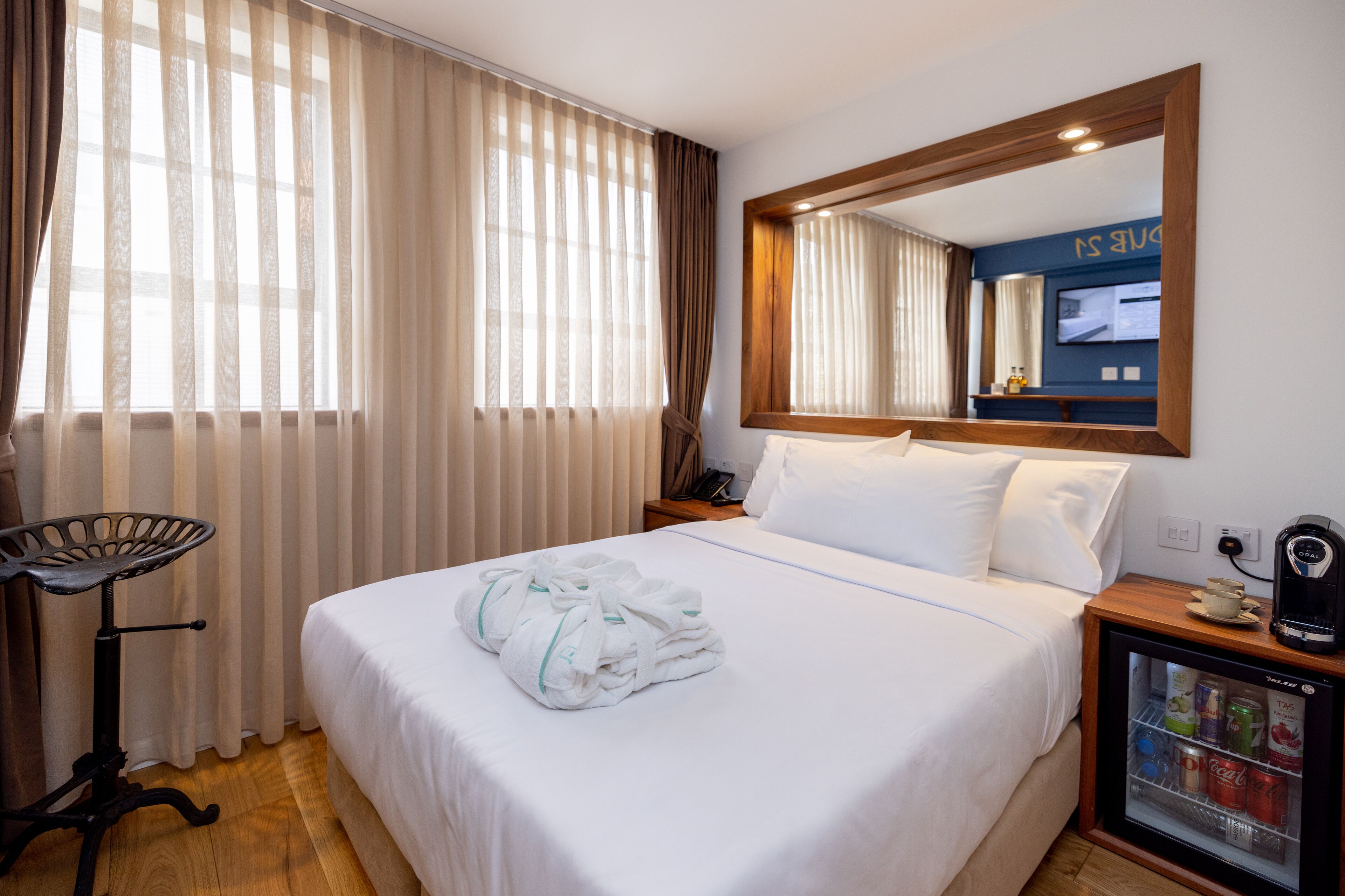 A hotel room with a neatly made bed, a small bedside table with a lamp, a minibar, and a large mirror on the wall.