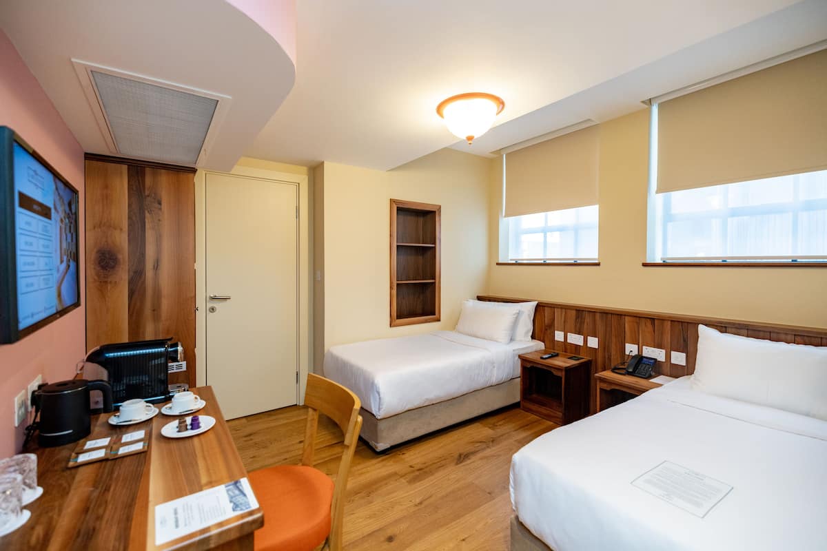 Une chambre d’hôtel avec deux lits, un bureau avec une machine à café, une chaise et une petite étagère.