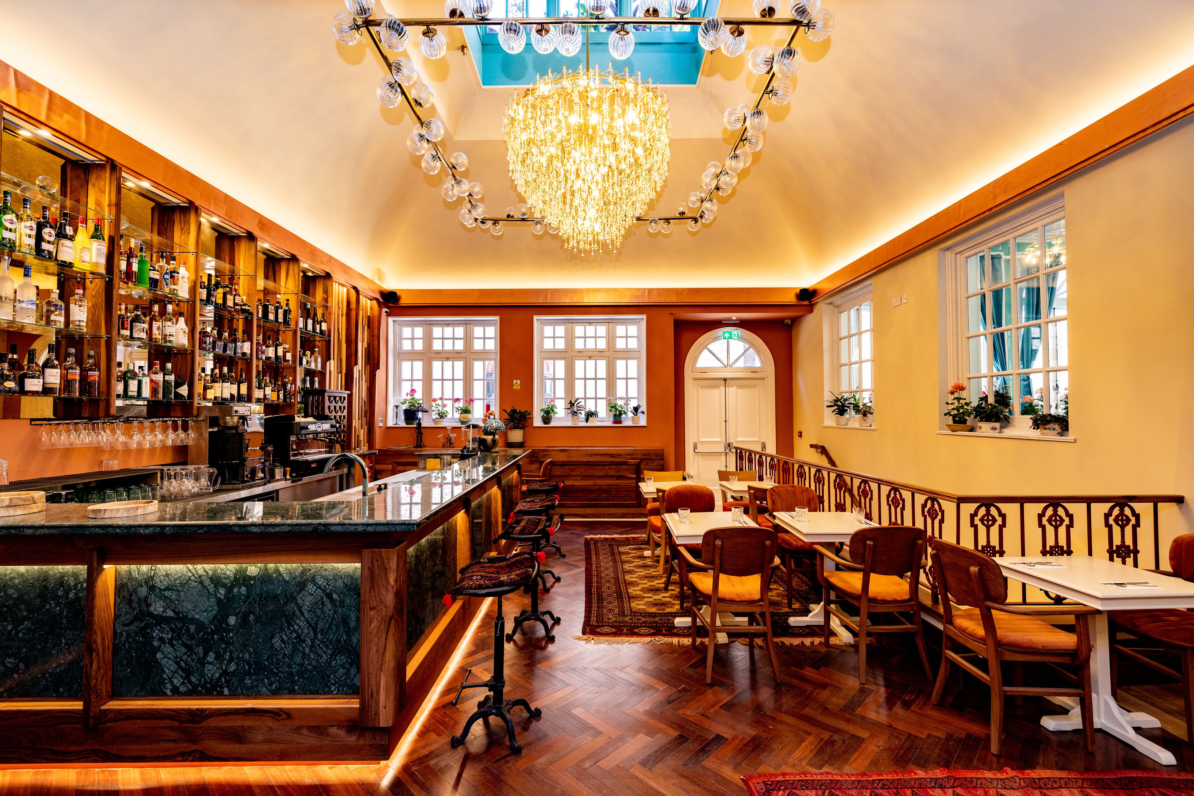 A bar area with a long counter, multiple stools, and a chandelier. There are wooden walls, a patterned rug, and a dining area with tables and chairs.