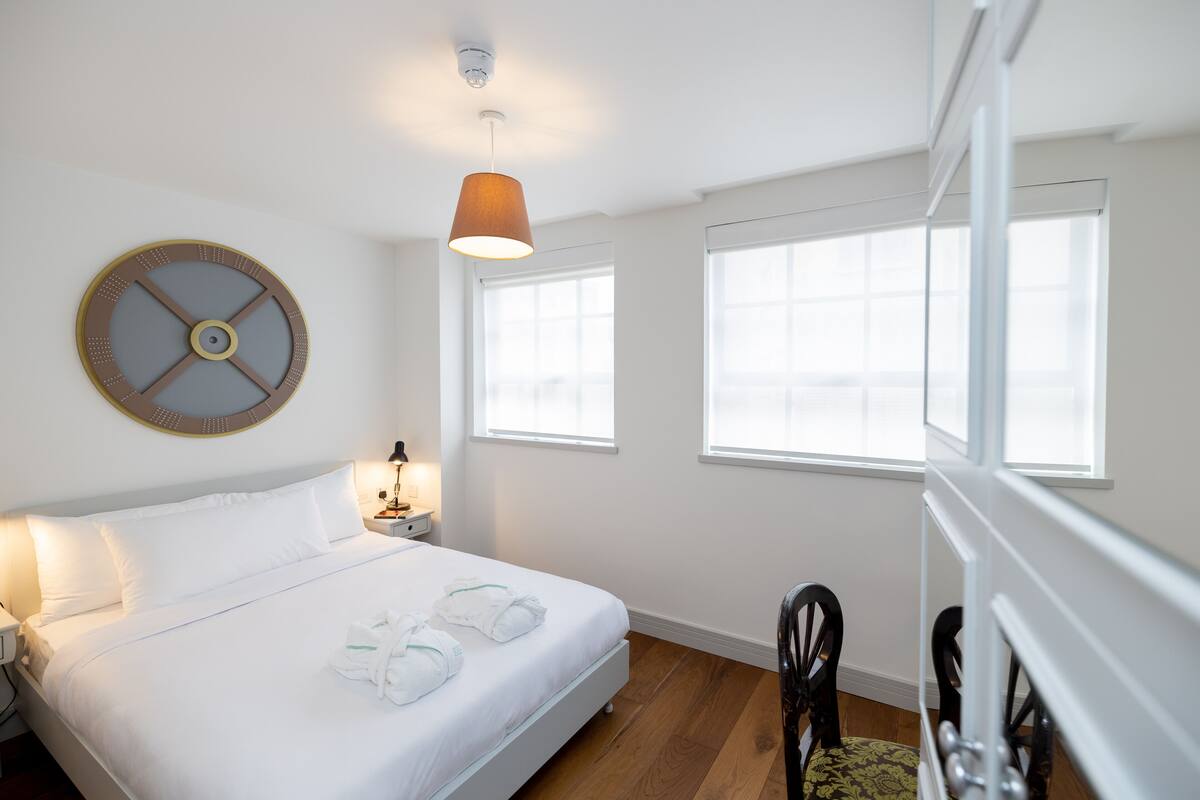 Une chambre d’hôtel avec un grand lit, deux tables de chevet, une horloge murale ronde, une lampe suspendue et une fenêtre donnant sur un beau paysage.