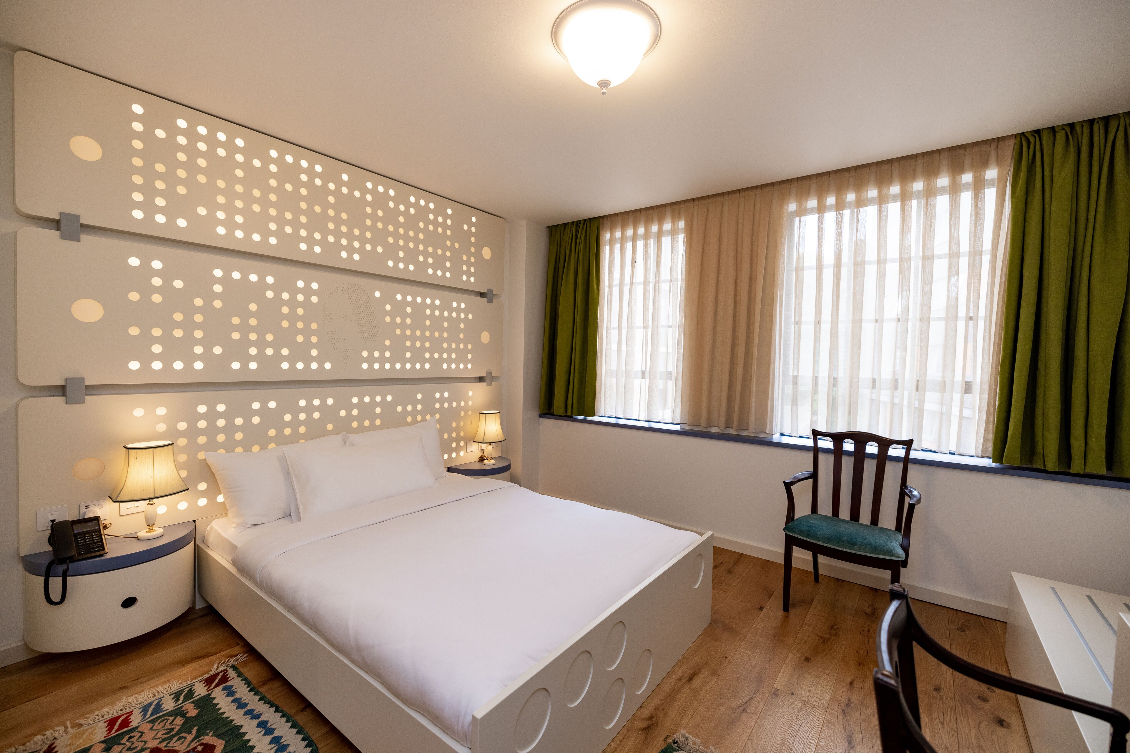Ein Schlafzimmer mit einem Bett, zwei Nachttischen mit Lampen, einem Sessel, einem Fenster mit Vorhängen und einem gemusterten Teppich.