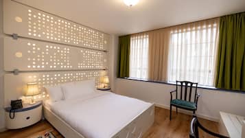 Ein Schlafzimmer mit einem Bett, zwei Nachttischen mit Lampen, einem Sessel, einem Fenster mit Vorhängen und einem gemusterten Teppich.