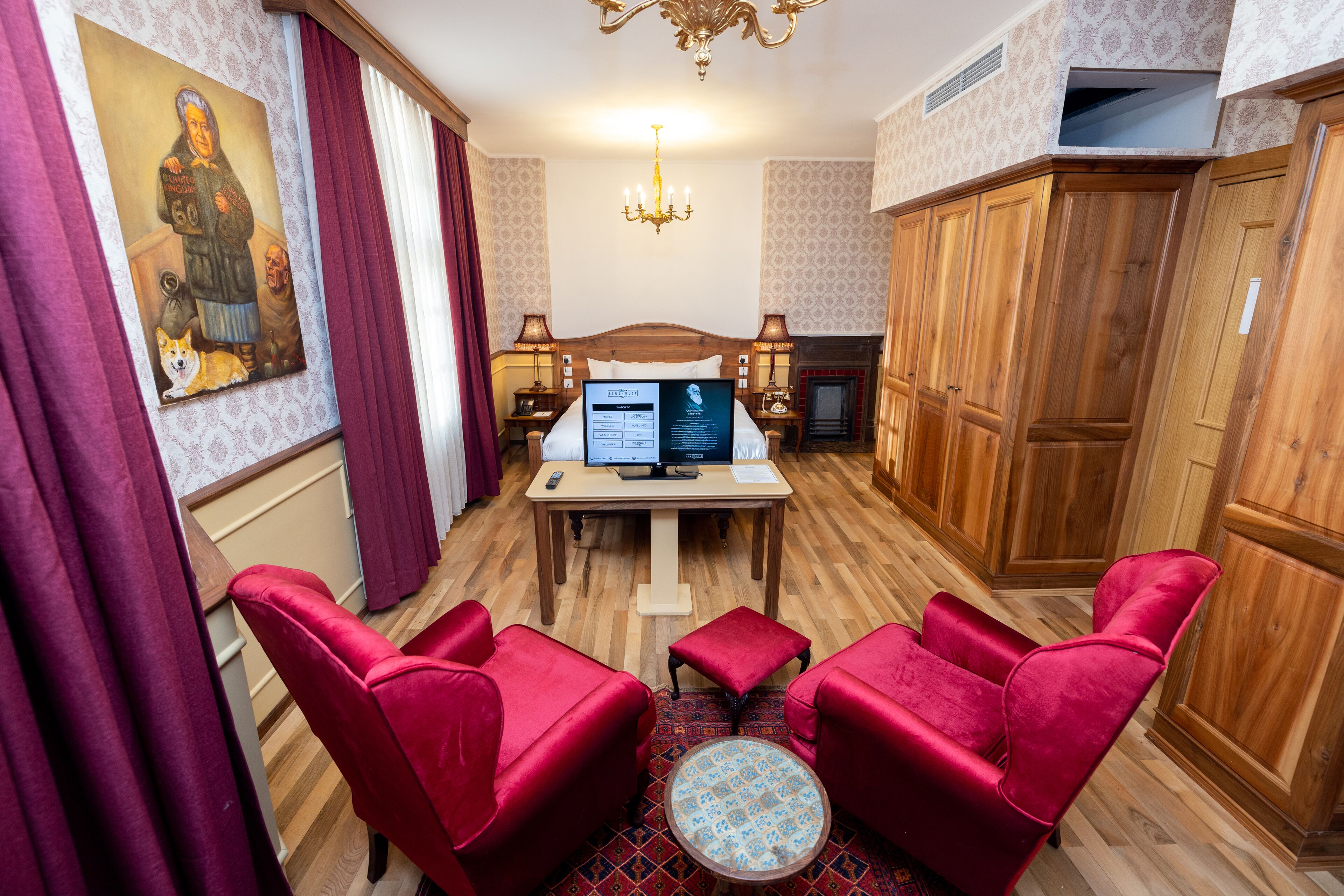Habitación de hotel con piso de madera, sillas tapizadas en rojo, una pequeña mesa redonda, un escritorio con computadora y papeles, un cuadro en la pared y una araña.