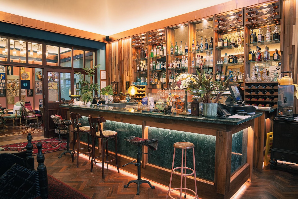 Hotellbar med grønn bardisk, barkrakker og hyller fulle av flasker i varmt lys.