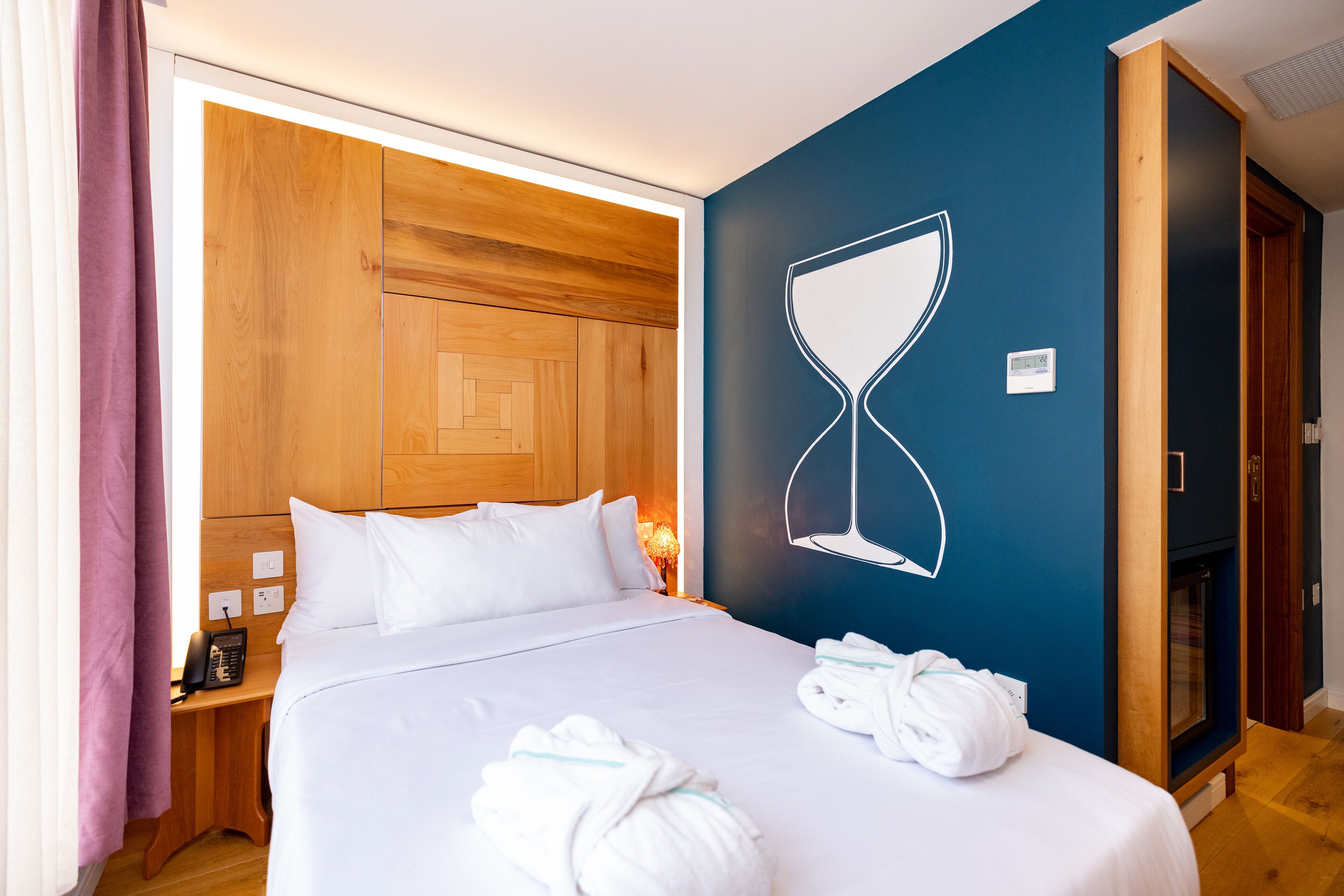 Een hotelkamer met een bed, houten hoofdeinde, een wandklok en een handdoekrek met handdoeken.