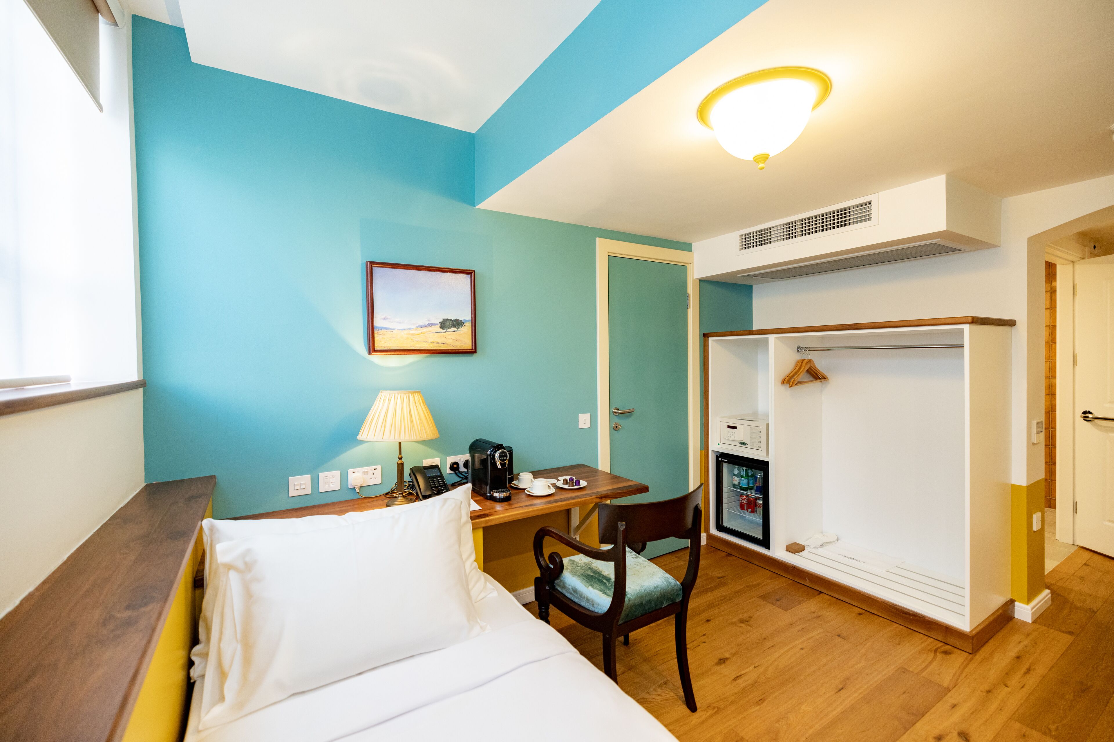 Una habitación de hotel con cama, escritorio con lámpara, silla, perchero y armario.