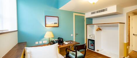 A hotel room with a bed, a desk with a lamp, a chair, a coat rack, and a wardrobe.