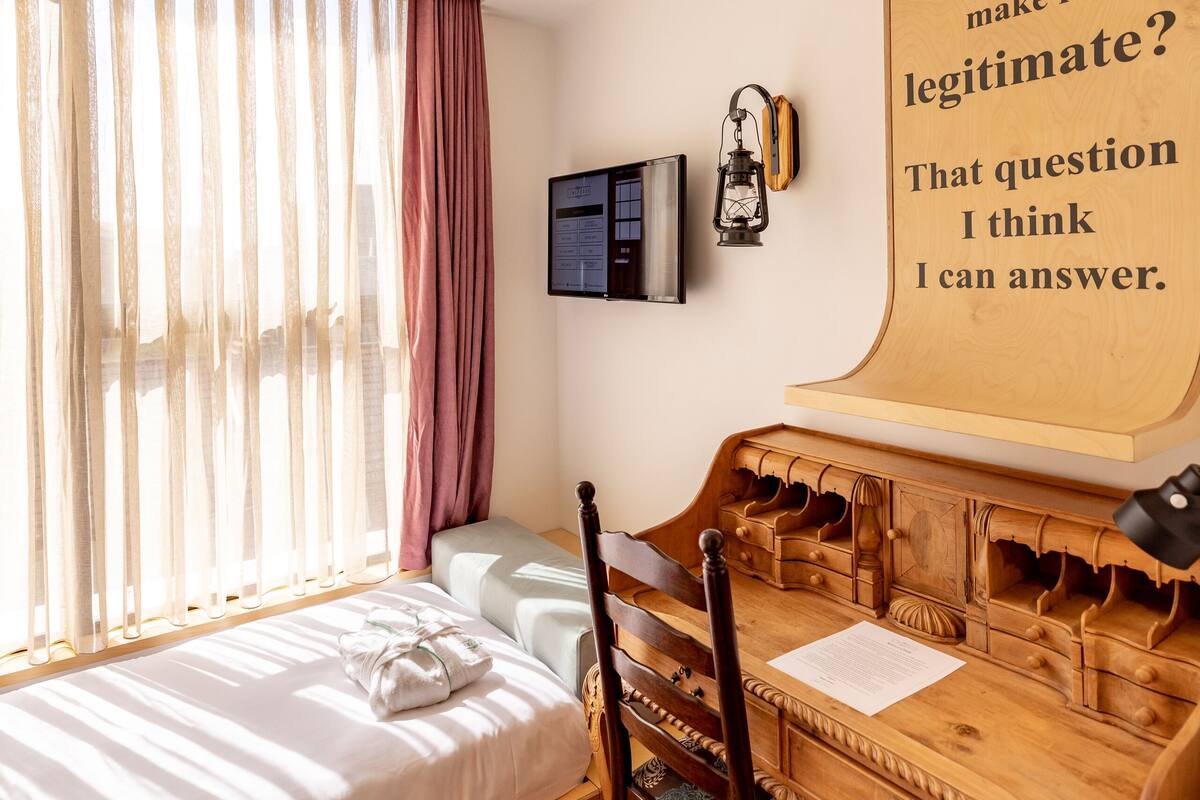 Une chambre à coucher avec un lit, un bureau en bois, une chaise et une lampe fixée au mur.