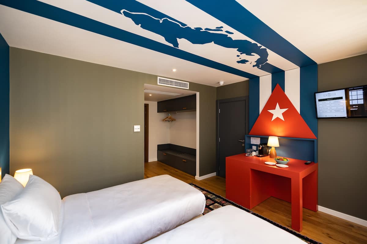Une chambre d’hôtel avec deux lits, un bureau rouge avec une lampe et une carte du monde peinte au plafond.