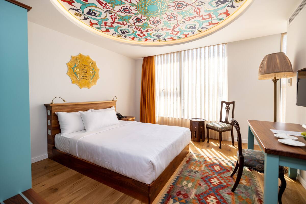 Une chambre d’hôtel avec un lit en bois, un bureau avec une lampe, une chaise, une télévision et un tapis à motifs.