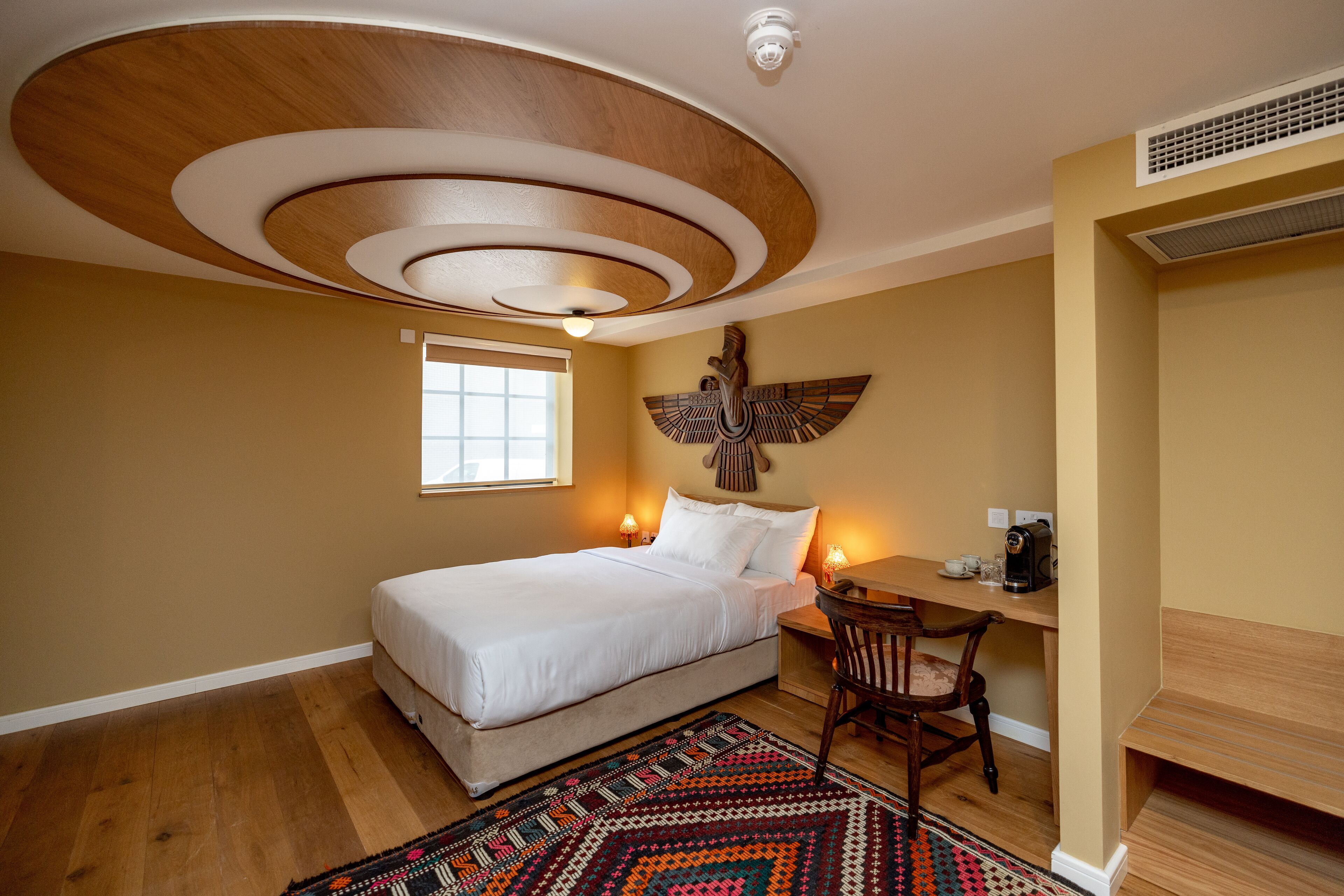 Ein Schlafzimmer mit einem Bett, einem Schreibtisch und einem Stuhl. Es gibt ein Fenster, ein an der Wand befestigtes Kunstwerk und eine Deckenleuchte.
