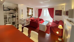Un salon moderne avec un canapé rouge, une table basse blanche et un tapis rouge. On y trouve une étagère blanche ornée d’objets décoratifs et un miroir. Une lampe rouge est posée sur le sol, et une table blanche se trouve au premier plan.