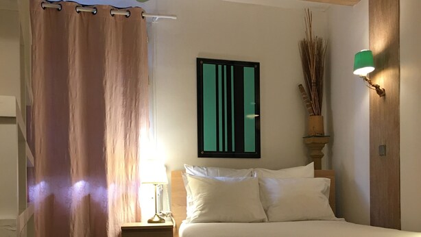 Une chambre à coucher avec un lit en bois, une table de chevet, un ventilateur de plafond vert, une lampe fixée au mur et une fenêtre avec des rideaux.