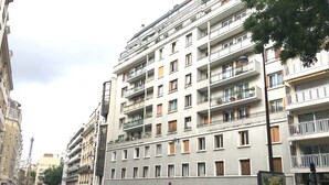 Un immeuble à plusieurs étages doté de balcons, donnant sur la rue, avec des voitures garées et une enseigne.