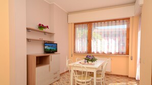 Ein Zimmer mit Fernseher auf einer Holzkommode, einem Tisch mit Stühlen, einem Fenster mit Vorhängen und einem gemusterten Boden.