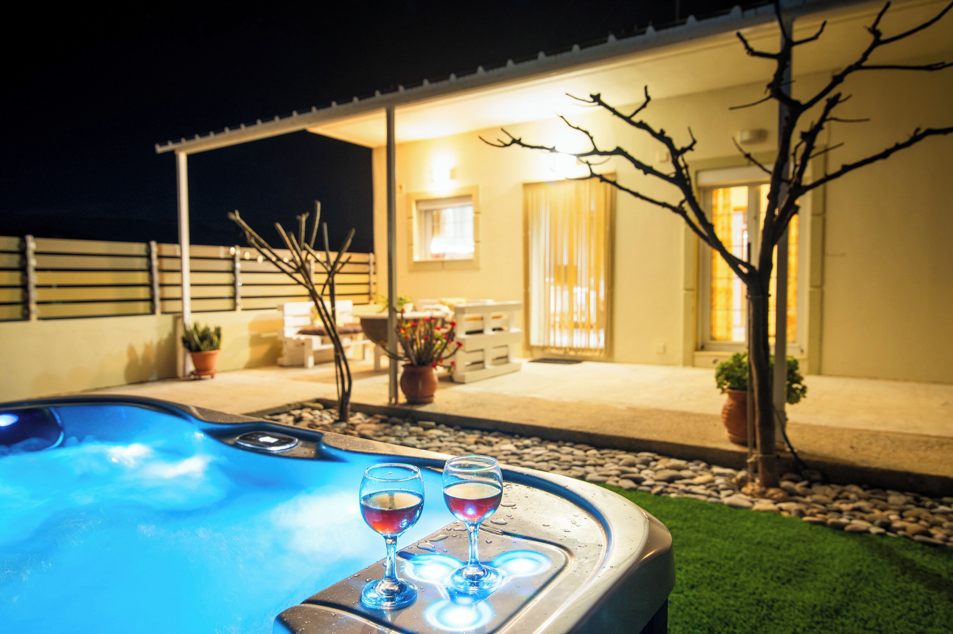 A hot tub with two glasses of red wine, surrounded by a patio area with potted plants and a building in the background.