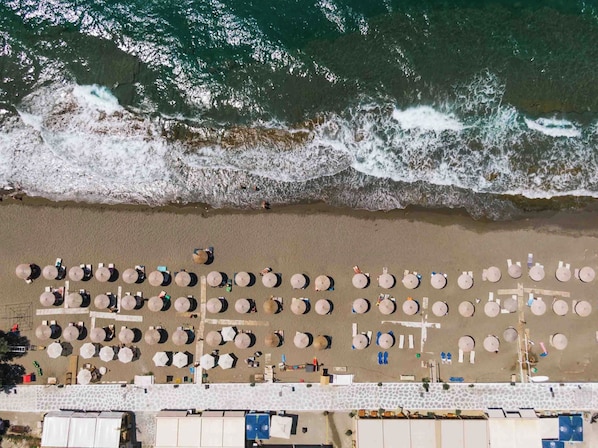 An aerial view of a beach with rows of round tables and umbrellas, a stone pathway, and a building with a flat roof.