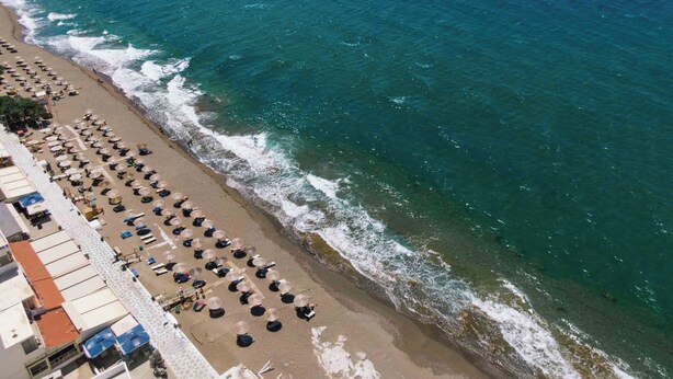 An aerial view of a beach with rows of sunbeds and umbrellas, a clear blue sea, and a building with a red roof on the left. - Family friendly holiday home  (Neo Kalamaki)